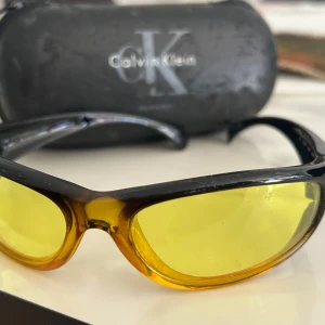 Gula solglasögon från Calvin Klein - Snygga gula solglasögon från Calvin Klein med en sportig look. De har en svart båge och gula linser som ger en cool kontrast. Perfekta för att sticka ut i mängden och skydda ögonen med stil.