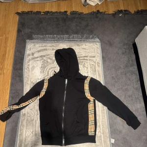 Svart hoodie med rutiga detaljer - Säljer en svart hoodie med dragkedja och klassiska rutiga detaljer längs ärmarna och sidorna. Perfekt för en stilren och bekväm look. Hoodien har en justerbar huva och fickor framtill.