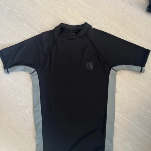 Solskyddströja Svart och grå  - Snygg solskyddströja. Svart med grå sidopaneler och korta ärmar. Perfekt för att skydda sig under soliga dagar. Emblem på bröstet har trillat bort i tvätt. Rök och djurfritt hem. 