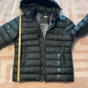 Svart pufferjacka från Boss - Säljer en svart pufferjacka från Boss med dragkedja och broderad logga. Jackan har långa ärmar och en huva för extra skydd. Perfekt för kyligare dagar. Jackan är splitterny men för liten för mig. Den gick sönder under ena armen därav priset. Se bild. 