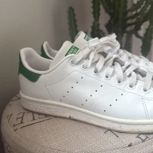 Vita Adidas Stan Smith sneakers - Klassiska vita Adidas Stan Smith sneakers med gröna detaljer. Skorna har snörning och en rund tå. Perfekta för en stilren och tidlös look.
