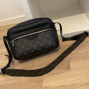 Svart axelväska med monogram - Snygg svart axelväska med klassiskt monogrammönster. Väskan har en justerbar axelrem och en praktisk dragkedja framtill. Perfekt för att bära dina dagliga nödvändigheter med stil.