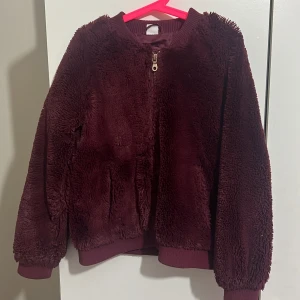 Vinröd teddyjacka med dragkedja - Mysig vinröd teddyjacka med dragkedja framtill. Jackan har långa ärmar och ribbade muddar vid ärmslut och nederkant. Perfekt för kyliga dagar.