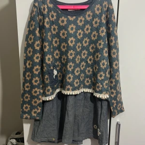 Blommig klänning med spetsdetaljer - Charmig klänning med blommigt mönster i beige och blått. Den har långa ärmar och en söt spetskant vid midjan. Kjoldelen är i en mörkare blå ton med dekorativa knappar.