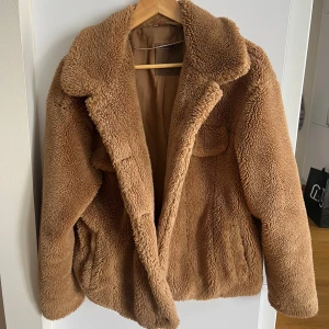 Brun teddyjacka - Mysig brun teddyjacka från Pull&Bear med långa ärmar och en klassisk krage. Perfekt för kyliga dagar när du vill hålla dig varm och stilren. Jackan har en mjuk och fluffig yta som ger en trendig look.