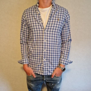 Blå och vit rutig skjorta från Ralph Lauren - Snygg blå och vit rutig skjorta från Ralph Lauren med klassisk krage och knappar framtill. Skjortan har långa ärmar och en liten broderad logga på bröstet. Perfekt för en stilren look. 