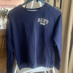 Blå sweatshirt från GANT - Säljer en stilren blå sweatshirt från GANT med tryck 'GANT 1949' på bröstet. Tröjan har långa ärmar och en rund halsringning. Perfekt för en avslappnad stil. 🌀