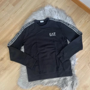 Svart långärmad tröja från Emporio Armani - Snygg svart långärmad tröja från Emporio Armani med EA7-logga på bröstet och längs ärmarna. Perfekt för en stilren look. Tröjan har en rund halsringning och är tillverkad i ett mjukt material.