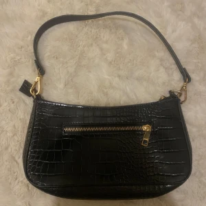 Svart axelväska i syntet med krokodilmönster - Snygg svart axelväska i syntet med krokodilmönster och guldfärgade detaljer. Väskan har en praktisk dragkedja framtill och en justerbar axelrem. Perfekt för att ge en stilren touch till din outfit.