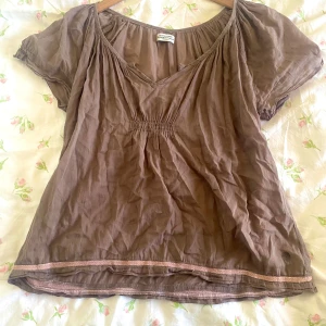 Brun blus från Hunkydory - Säljer en brun blus från Hunkydory Essentials med korta ärmar och v-ringning. Blusen har fina detaljer med resår i midjan och spetskant vid ärmslut och nederkant. Perfekt för en avslappnad stil.