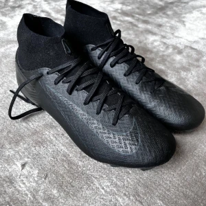 Svarta fotbollsskor från Nike - Säljer ett par svarta Nike Mercurial fotbollsskor med snörning och hög ankelkrage. Skorna har en strömlinjeformad design med rund tå och är perfekta för spel på gräs. De är tillverkade i syntetmaterial för optimal prestanda på planen.
