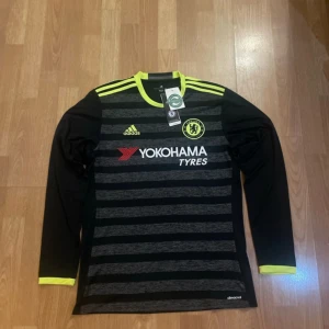 Svart och grå Chelsea fotbollströja från Adidas - Säljer en svart och grå randig Chelsea fotbollströja från Adidas med långärmad design. Tröjan har gula detaljer och klubbens logga på bröstet. Perfekt för fans som vill visa sitt stöd. Tillverkad av återvunnen polyester. Priset kan dissekeras!!