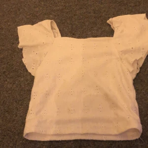 Vit blus från Kappahl - Säljer en söt vit blus från Kappahl med korta ärmar och ett diskret broderat mönster. Perfekt för varma dagar med sin luftiga design. Blusen har en klassisk passform och är tillverkad i ett mjukt material.