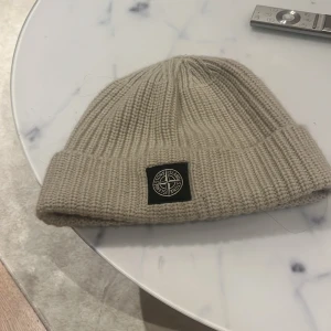 Beige stickad mössa från Stone Island - Snygg beige stickad mössa från Stone Island med ribbad design och uppvikt kant. Perfekt för att hålla värmen under kyliga dagar. Märkeslogga framtill ger en stilren touch.