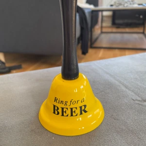 Gul klocka med texten 'Ring for a Beer' - En rolig gul klocka med texten 'Ring for a Beer' i svart. Perfekt för att liva upp festen eller som en skojig present. Klockan har ett svart handtag och är lätt att använda.