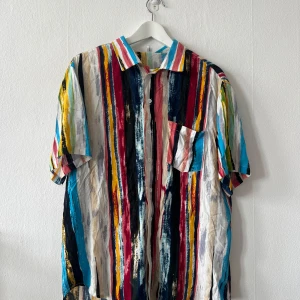 Flerfärgad randig skjorta - Säljer en färgglad skjorta med vertikala ränder i blått, rött, gult, svart och vitt. Skjortan har korta ärmar och en klassisk krage. Perfekt för en somrig look!