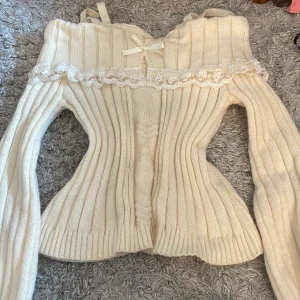 Vit ribbad offshoulder tröja med spetsdetaljer - Säljer en söt vit ribbad offshoulder tröja med spetsdetaljer och en liten rosett framtill. Tröjan har långa ärmar och en figurnära passform. Perfekt för en romantisk look!