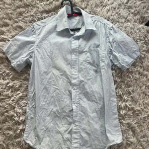 Vit och blå randig skjorta - Snygg vit och blå randig skjorta med korta ärmar och klassisk krage. Perfekt för en avslappnad men stilren look. Skjortan har en bröstficka och knappar framtill.