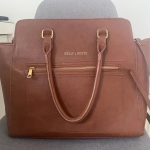 Brun handväska  - Snygg brun handväska med guldiga detaljer från Holly & White i skinn med praktisk dragkedja framtill. Väskan har både handtag och en justerbar axelrem för flexibilitet. Jätte fint skick