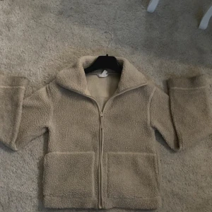 Beige teddyjacka från H&M - Mysig beige teddyjacka med dragkedja framtill. Jackan har en hög krage och långa ärmar, perfekt för kyligare dagar. Den har en avslappnad passform och är gjord i ett mjukt teddy-material.
