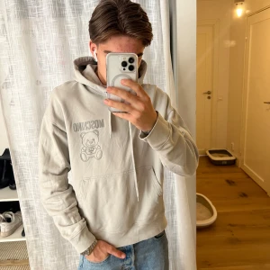 Beige hoodie från Moschino - Säljer en snygg beige hoodie från Moschino med ett coolt nalletryck på framsidan. Hoodien har en klassisk passform med långa ärmar och en praktisk ficka framtill. Perfekt för en avslappnad stil!