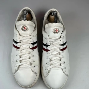 Vita sneakers från moncler  - Snygga vita sneakers med klassiska ränder i svart och rött på sidan. Skorna har snörning och en rund tå, vilket ger en stilren look. Perfekta för en avslappnad stil.