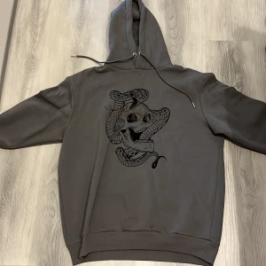 Grå hoodie med dödskalle och orm - Säljer en grå hoodie från MAN med en cool design av en dödskalle och orm på framsidan. Hoodien har en justerbar huva med dragsko och ribbade muddar vid ärmslut och nederkant. Perfekt för en avslappnad stil.