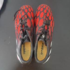 Adidas Predator fotbollsskor röd/svart - Snygga Adidas Predator fotbollsskor med röd och svart ovandel, vita ränder på sidorna och orange dobbar. Skorna har snörning och ett mönster med röda hexagoner. Perfekta för dig som vill sticka ut på planen.
