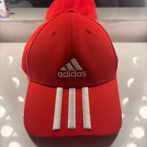 Röd Adidas keps med vita ränder - Snygg röd keps från Adidas med klassiska tre vita ränder framtill och broderad logga. Justerbar rem baktill med Adidas-detalj. Den är i fint skick, någon enstaka defekt men ser annars väldigt bra ut. Vid frågor är det bara att höra av sig!
