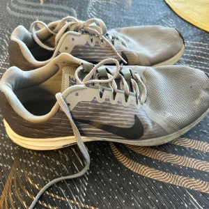 Grå Nike sneakers med mesh och snörning - Snygga grå Nike sneakers med svart swoosh på sidan. Ovandelen är i mesh för bra andningsförmåga och skorna har klassisk snörning. Sulan är vit med svart mönstrad undersida. Perfekta för träning eller vardagsstil.