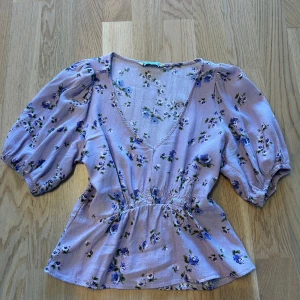 Ljuslila blommig blus från Stradivarius - Supersöt ljuslila blus från Stradivarius med blått blommönster, v-ringning och puffiga korta ärmar. Smockad midja ger en snygg siluett!