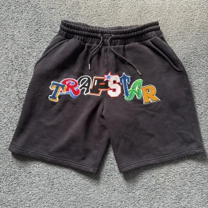 Unika svarta Trapstar shorts - Svarta shorts från Trapstar med färgglad broderad logga framtill. Mjuk bomullskänsla, elastisk midja med snörning och två sidofickor. Perfekta för chill dagar eller streetwear-stil. Snygg kontrast mellan svart tyg och broderade bokstäver i blått, rött, grönt, gult och vitt.