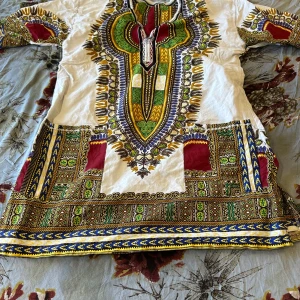 Färgstark afrikansk henley-skjorta - Unik henley-skjorta med färgstarkt afrikanskt mönster i vitt, grönt, gult, blått och rött. Skjortan har lång ärm, v-ringning med snörning och detaljerade tryck längs kanter och bröst. Perfekt för dig som vill sticka ut med en riktigt cool och färgglad look.