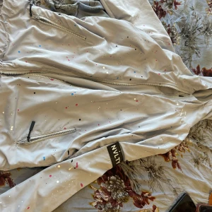 Ljusgrå vindjacka från NVLTY - Snygg ljusgrå vindjacka från NVLTY med färgstänk i rosa, blått, svart och vitt. Jackan har dragkedjor framtill och på fickorna samt en cool patch på ärmen. Perfekt för dig som gillar streetstyle och vill sticka ut lite extra.