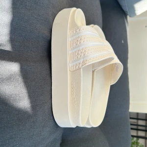 Vita Adidas sandaler med platåsula - Snygga vita Adidas sandaler med platåsula och klassiska tre ränder över foten. Tillverkade i syntetmaterial med strukturerad ovandel och präglad Adidas-logga på sidan. Perfekta för sommardagar och chill vibes.