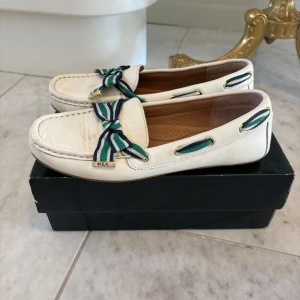 Vita loafers från Lauren Ralph Lauren - Snygga vita loafers från Lauren Ralph Lauren i skinn med klassisk seglarsko-look. Dekorativa randiga band i blått, grönt och vitt med metall-detaljer pryder ovandelen. Perfekta för en preppy stil.