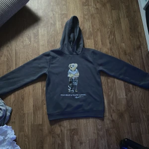 Mörkblå Polo Bear hoodie från Ralph Lauren - Snygg mörkblå hoodie från Ralph Lauren med ikoniska Polo Bear-trycket på bröstet. Tröjan har huva, långa ärmar och en klassisk passform. Perfekt för dig som gillar streetstyle och vill sticka ut med en cool detalj på din outfit. Finns ingen tag i nacken därav priset 