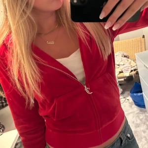 Röd velourjacka från Juicy Couture - Säljer en ikonisk röd velourjacka från Juicy Couture med dragkedja och huva. Jackan har långa ärmar, ribbade muddar och två stora fickor framtill. Perfekt för en chill och trendig look, klassisk Y2K-vibe.