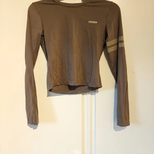 Brun långärmad träningströja Stronger - Sportig brun långärmad träningströja från Stronger med två reflekterande ränder på ena ärmen och små hål i tyget på ryggen för extra ventilation. Tillverkad i stretchigt material som sitter skönt på kroppen. Perfekt för dig som gillar att träna med stil.