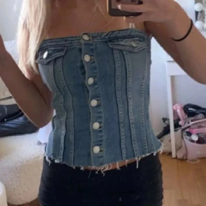 Jeans korsett-topp med knappar från Zara - Säljer en snygg axelbandslös korsett-topp i blå denim med silverfärgade knappar framtill och rå kant nertill. Toppen har två dekorativa fickor upptill och markerade sömmar som ger en cool och trendig look. Perfekt för dig som vill sticka ut i sommar! St L men sitter som en M/S då den är tajt pga denim materialet