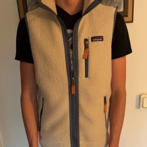  Patagonia väst - Säljer en beige fleeceväst från Patagonia med blå dragkedjor och snygga läderdetaljer på zipparna. Västen har hög krage och tre fickor med dragkedja. Perfekt att slänga över en hoodie eller t-shirt för extra värme.