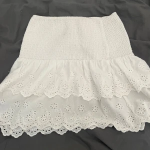 Vit volangkjol med broderade spetsdetaljer - Supersöt vit kortkjol med bred smock i midjan och dubbla volanger. Kjolen har broderade blommor och spetskant som ger en romantisk vibe. Perfekt för varma dagar och sommarhäng.