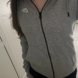 Grå Nike hoodie med dragkedja S - Säljer en grå Nike hoodie i storlek S med klassisk vit logga på bröstet. Tröjan har huva med snörning, hel dragkedja och två fickor framtill. Perfekt för chill dagar eller när du vill ha en sportig look.