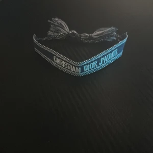 Blått Christian Dior J'Adior armband - Oanvänd.Snyggt blått armband från Christian Dior med texten 'J'Adior' broderad i vitt. Armbandet har justerbara tofsar i ändarna och är gjort i ett mjukt tygmaterial. Perfekt accessoar för att lyfta din outfit med en lyxig touch.