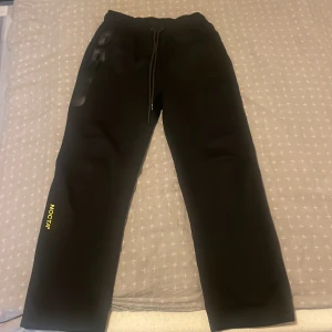 Svarta NOCTA mjukisbyxor med snörning - Svarta mjukisbyxor från NOCTA med gul logga på benet och snörning i midjan. Byxorna har en clean look och raka ben, perfekta för en chill och sportig stil.