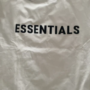 Vit Essentials t-shirt Fear of God - Vit t-shirt från Fear of God Essentials med svart tryck på bröstet. Klassisk rund halsringning och korta ärmar. Tillverkad i mjuk bomull för en clean och stilren look. Perfekt för dig som gillar minimalistisk streetwear.