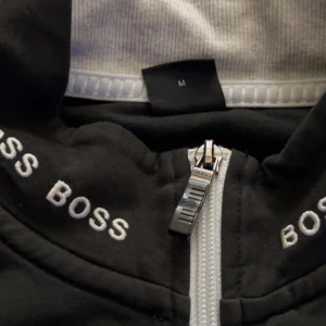 Svart bomberjacka från Hugo Boss M - Svart bomberjacka från Hugo Boss med vit dragkedja och broderad logga på bröstet. Kragen har BOSS-tryck runtom för en clean och modern look. Jackan är i ett mjukt material och har långa ärmar. Perfekt för dig som gillar stilrena och sportiga vibes.