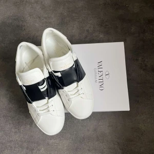 Valentino Garavani Open sneakers vit/svart - Snygga Valentino Garavani Open sneakers i vitt skinn med bred svart läderdetalj över vristen. Klassisk rund tå, vita snören och diskret logga på plösen. Rena linjer och chunky sula ger en modern vibe. Perfekta för dig som gillar exklusiva och stilrena sneakers. Använda fåtalgånger. Säljs pågrund av att dom var för så för mig men annars var dom riktigt sköna skor.