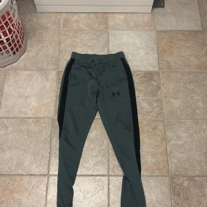 Under Armour träningsbyxor grå/svarta - Snygga träningsbyxor från Under Armour i mörkgrått med svarta sidopaneler. Byxorna har snörning i midjan och smal passform. Perfekta för gymmet eller löprundan, med sportig look och bekvämt material.
