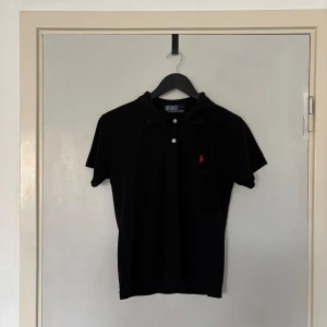 Svart pikétröja från Ralph Lauren - Klassisk svart pikétröja från Ralph Lauren med röd broderad logga på bröstet. Tröjan har korta ärmar, krage och två knappar framtill. Tillverkad i mjuk bomull som är skön mot huden. Perfekt för en clean och tidlös stil.